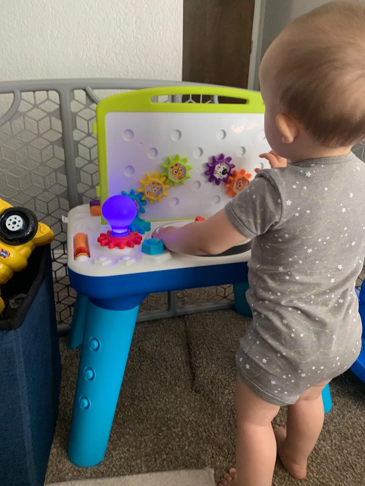 A standing infant works the gears and reaches for the glowing bulb at standing height.