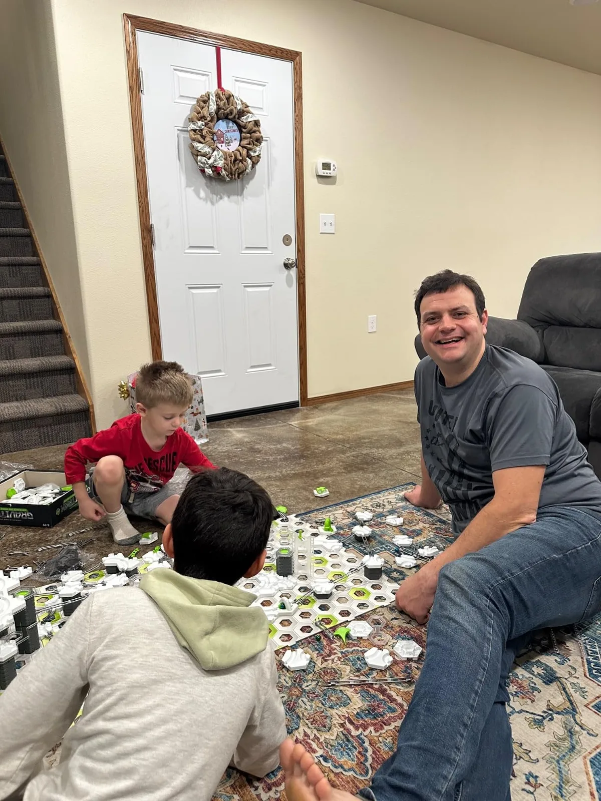 A father and two children deep in a build session, hex tiles spread across a living-room rug.