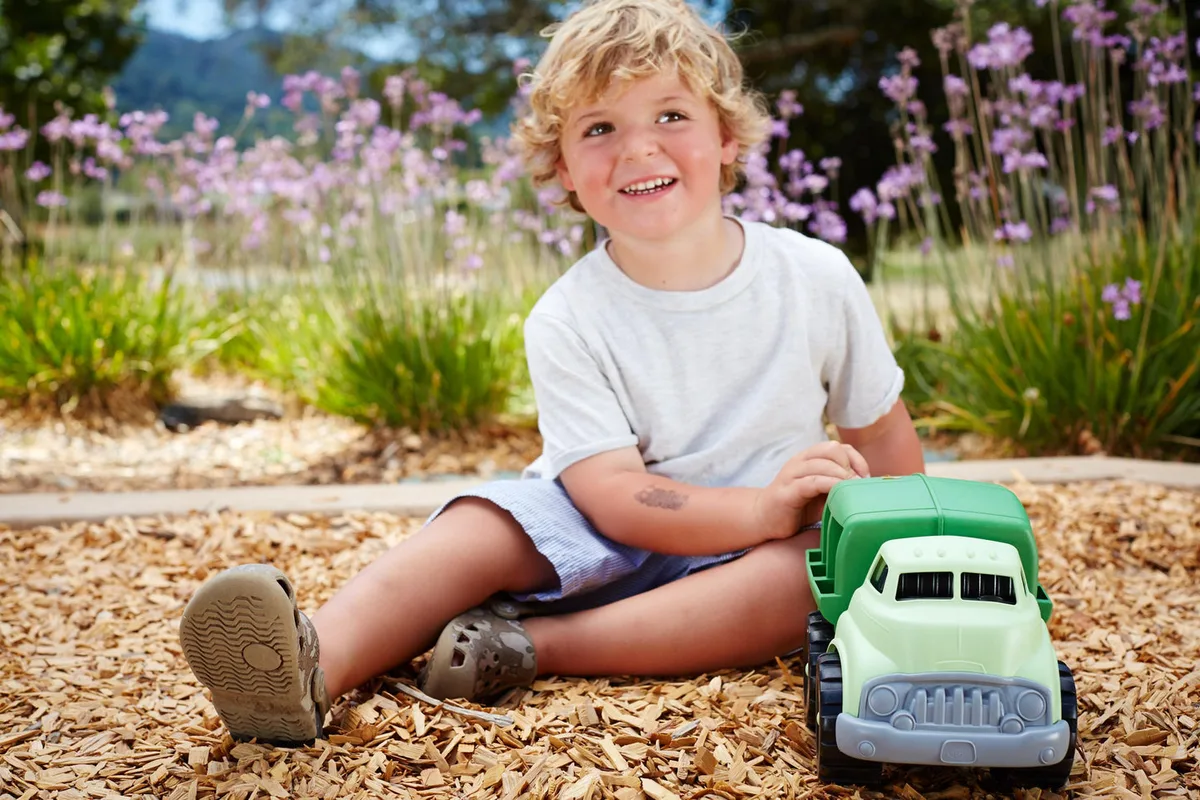 A young child plays outdoors with the recycling truck on wood chips beneath flowering plants.