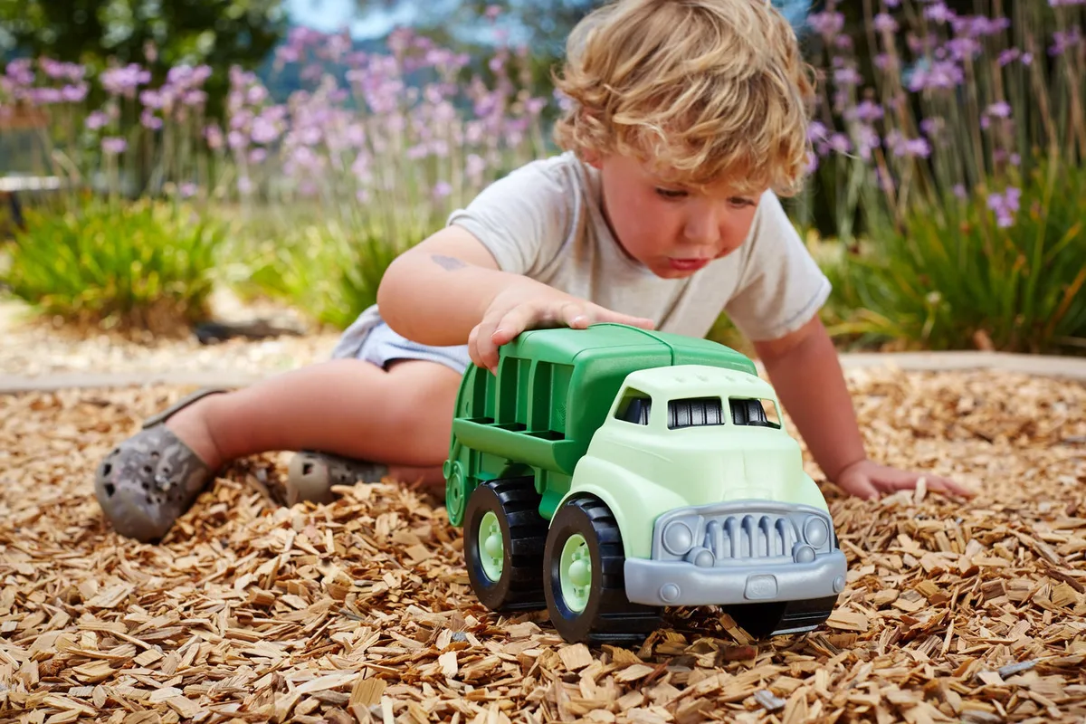 A toddler kneels on mulch, fully absorbed in pushing the recycling truck through outdoor play.