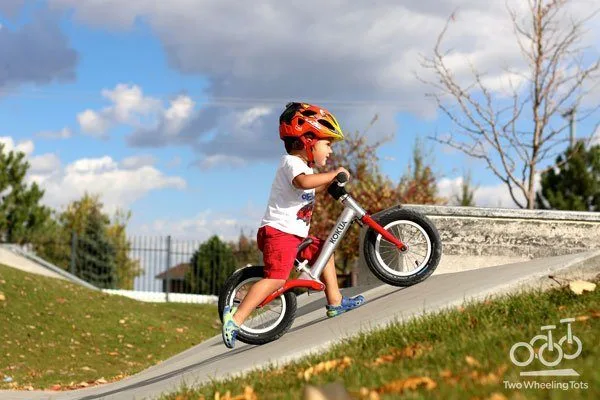 A toddler navigates a paved skate park ramp on the red KoKua Jumper balance bike.