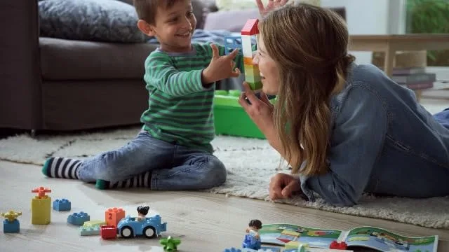 A parent and toddler build together on the rug, bricks scattered between them.