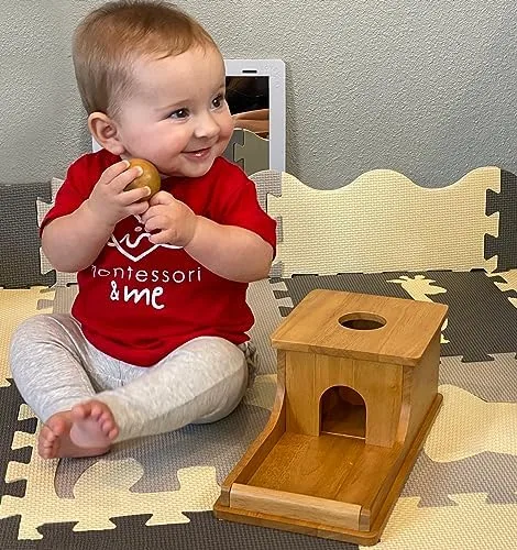 A baby pauses with the wooden ball, mid-experiment, the box waiting beside her.