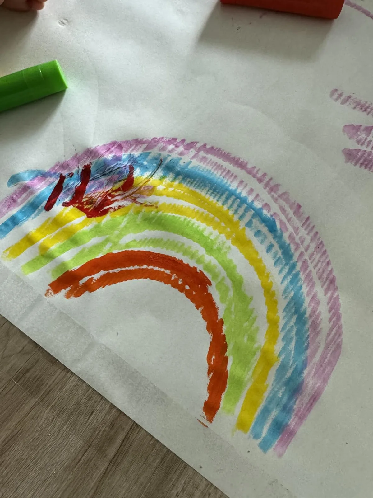 A toddler's rainbow streaks across paper, the green stick still uncapped beside the work.