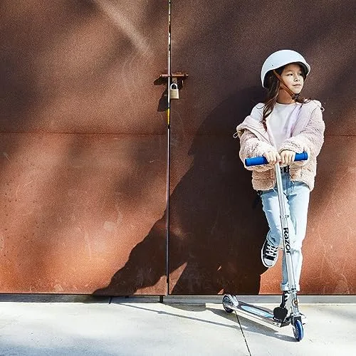 A rider pauses against a sunlit wall, helmet on, the aluminum frame visible underfoot.