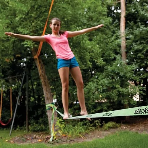 A child mid-traverse, arms wide — the slackline visibly bows under load between two trees.