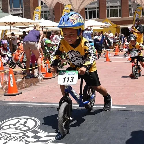 A young rider crosses the line at a Strider Cup heat, helmet on, both feet pushing.
