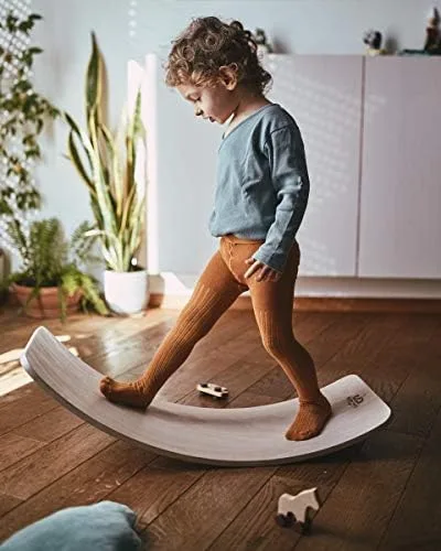 A toddler tests the wooden board's curve, balancing in a sunlit room with houseplants and white cabi